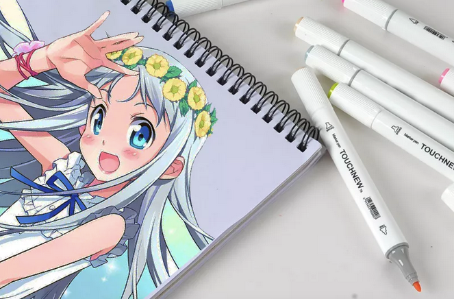 Several marker pens are scattered next to a sketchbook with a colourful manga illustration of a smiling girl. The cap is off on one pen, showing the fine nib.