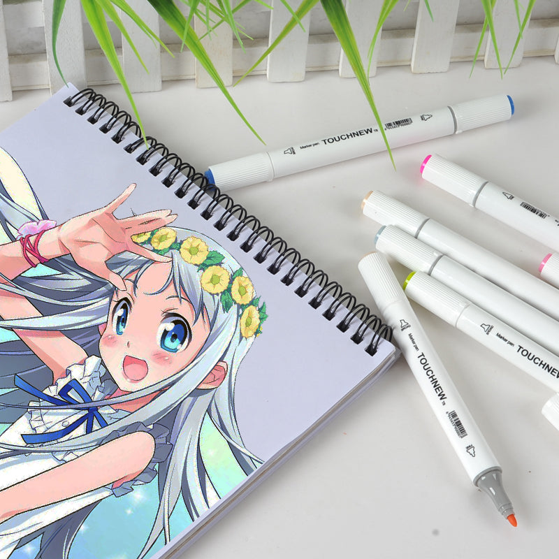 A handful of cylindrical alcohol markers are scattered on a surface next to a sketchbook with a manga illustration on it. One of the markers is uncapped, showing off the fine nib.