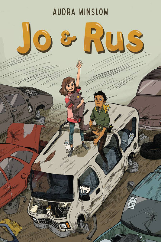 A young white girl with shoulderlength brown hair stands on top of a junked up wheel-less van, holding one arm in the air while the other holds a large one-eyed cat to her chest. A young man with short dark hair and goatee and tan skin sits next to her on the van roof, several smaller cats around him. In the background are several other scrap vehicles.